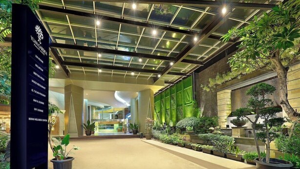 A modern hotel lobby with a glass ceiling, potted plants, and a sign listing amenities like a ballroom and wellness center. - Yuan Garden Pasar Baru (Jakarta)