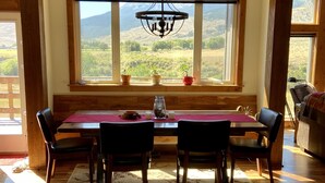 Un comedor con una mesa de madera, cuatro sillas, una araña y ventanas amplias con vistas a un paisaje montañoso.