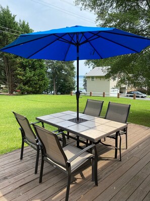 Eine Terrasse mit Tisch und Stühlen, einem blauen Sonnenschirm und Blick auf ein Haus und Bäume.