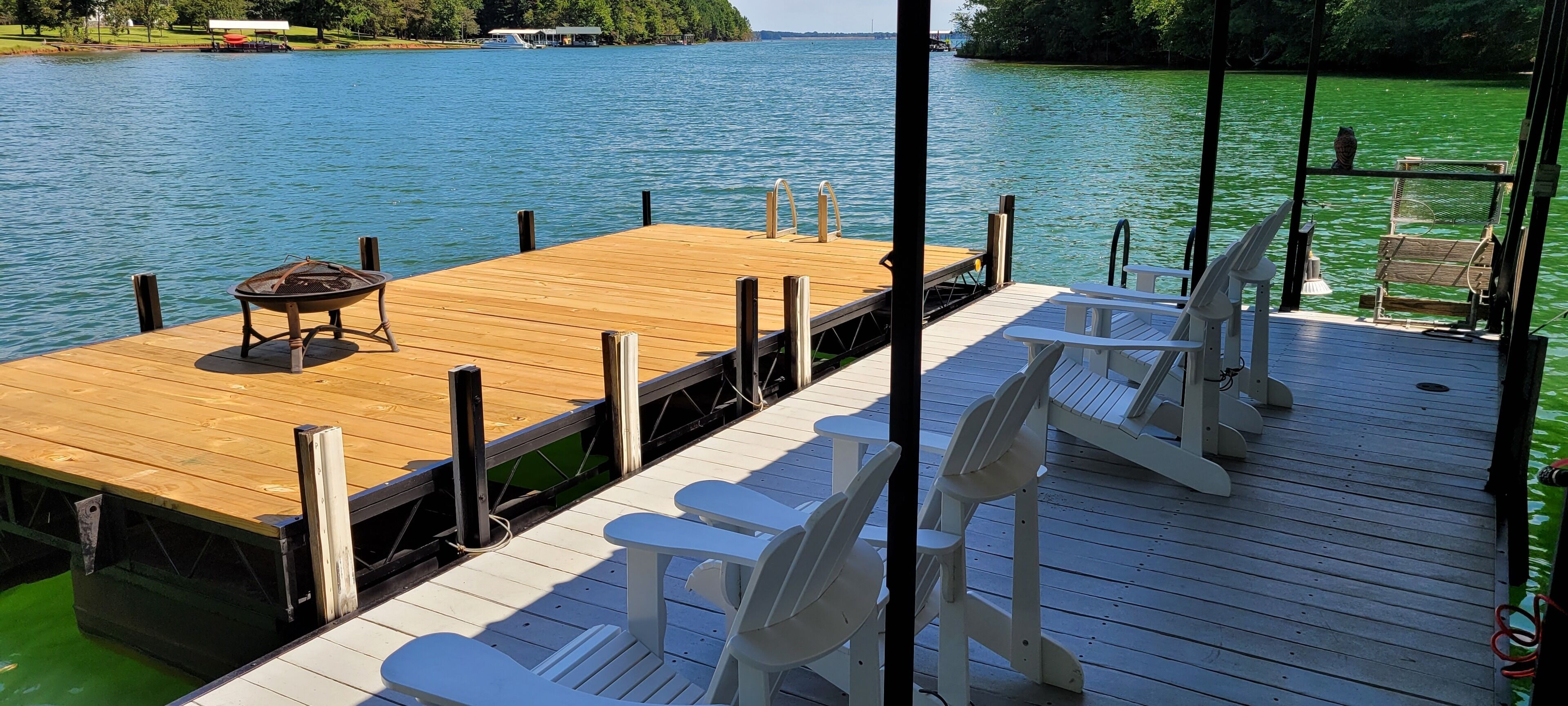 A wooden dock with white chairs and a table, overlooking a calm lake with a boat docked in the background.