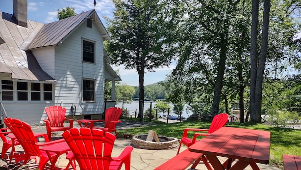 A patio with red Adirondack chairs and a wooden table, overlooking a lake and a white house.