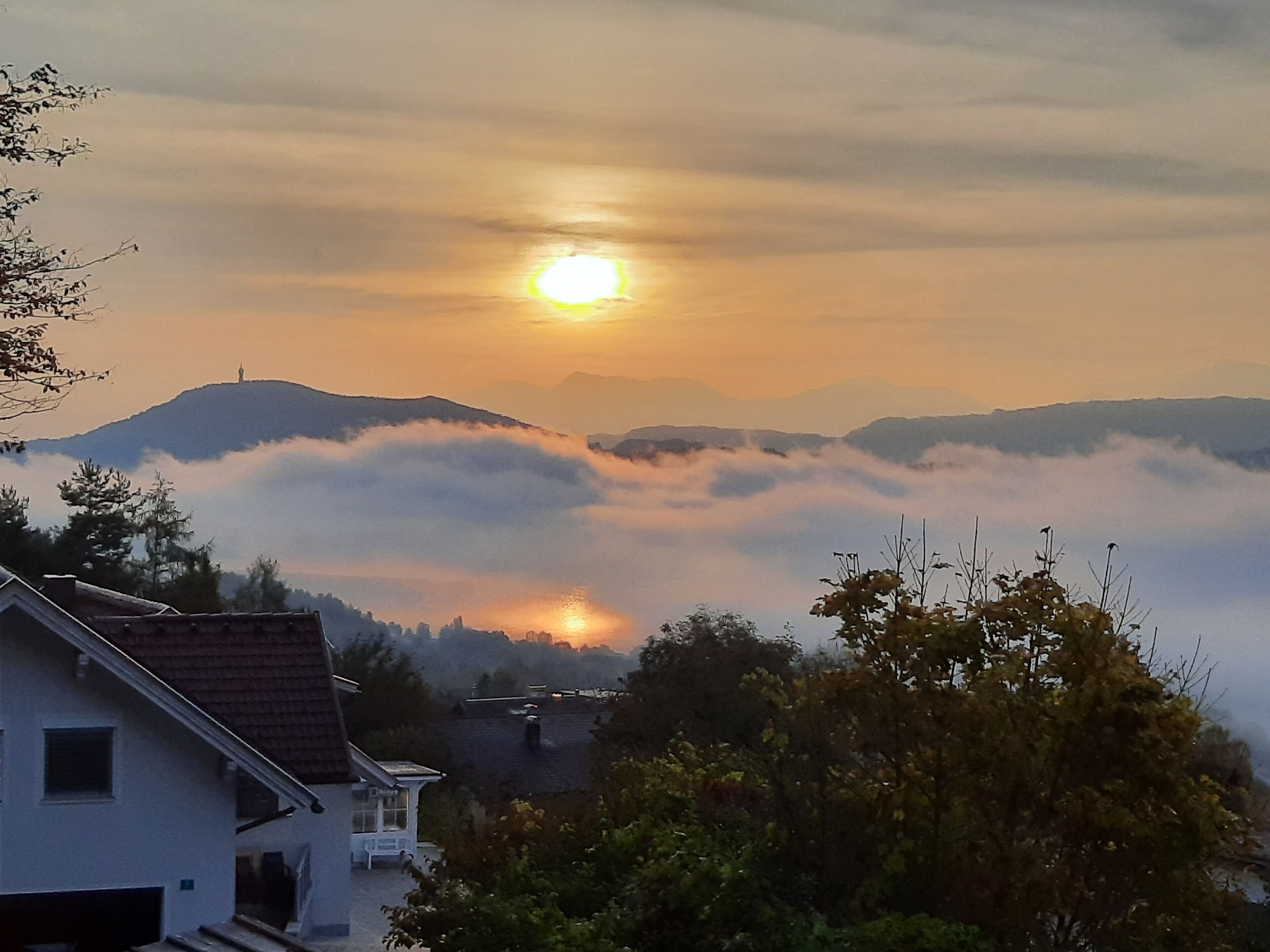 A house with a sloped roof, a small porch, and a view of the sun setting behind a mountain range partially covered by clouds.
