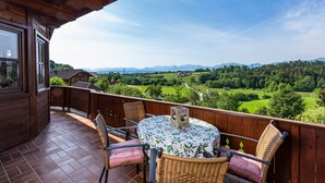 A wooden balcony with a table and chairs, overlooking a green landscape with mountains in the distance.