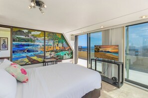 A hotel room with a large bed, a painting of a tropical landscape, a television, and a balcony with a view of the ocean.