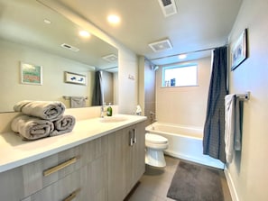 A modern bathroom with a sink, toilet, and bathtub. There are towels on the counter and a window above the bathtub.