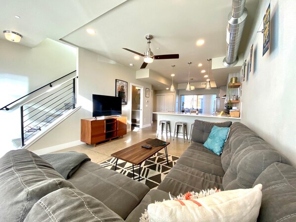 A modern living room with a gray sectional sofa, a wooden coffee table, and a flat-screen TV on a wooden stand. There is a staircase with a metal railing leading to an upper level.