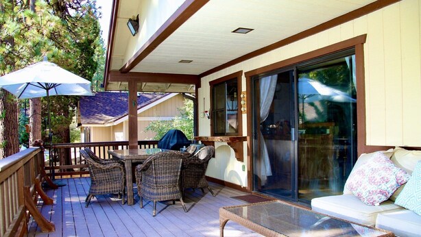 A covered outdoor patio with wicker furniture, a glass table, and a sliding glass door leading to a living area. - The Bear Slumbers in Big Bear, spa, WiFi, foosball, pool/ping-pong (Big Bear)
