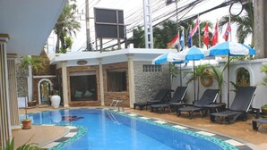 Une piscine entourée de chaises longues et de parasols, bordée de palmiers et d’un mur de pierre.