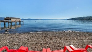 Una playa de guijarros con un muelle de madera y un edificio pequeño, con vistas a aguas tranquilas y colinas lejanas.