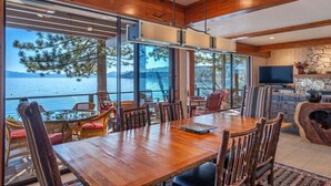 Un comedor amplio con mesa y sillas de madera, ventanas grandes con vista al agua y las montañas, y una chimenea de piedra.