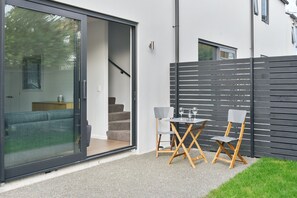 A modern outdoor patio with a sliding glass door leading to a living area, a small table with two chairs, and a black fence.