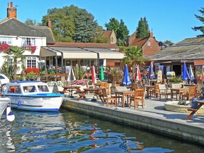 Een café aan de rivier met terras, een boot afgemeerd in de buurt, en een gebouw met een bordje 'AMUSEMENTS'.