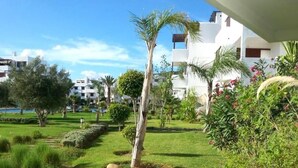 A well-maintained garden with a variety of plants and a palm tree, surrounded by modern buildings. - Apartment with pool view at La Cassia 4187 complex (Cabo Negro)
