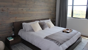 Una camera da letto moderna con una testiera in legno, un letto grande con biancheria da letto bianca, un comodino con un libro e una finestra con vista sugli alberi.