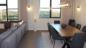 Un soggiorno moderno con divano, tavolo da pranzo e sedie, ampie finestre che si affacciano su una zona boschiva.