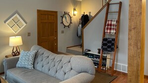 Ein Wohnzimmer mit grauem Sofa, einer Holztür, einer Wanduhr, einem Kleiderständer und einer Holztreppe mit Geländer.