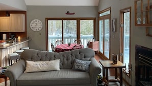 Ein gemütliches Wohnzimmer mit grauem Sofa, einem Holztisch und einem Kamin. Es gibt einen Essbereich mit rotem Tisch und Stühlen sowie eine Küche mit weißen Schränken und einer Uhr.