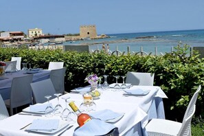A dining table set with white tablecloths, wine glasses, and a bottle, overlooking a coastal view with a historic building and greenery. - Marco's terrace with sea view - beach 50 m - promenade 50 m (Carovigno)