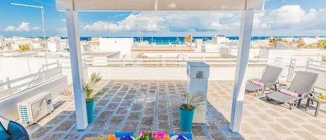 A rooftop terrace with a view of the sea, tables set with plates and drinks, and lounge chairs.
