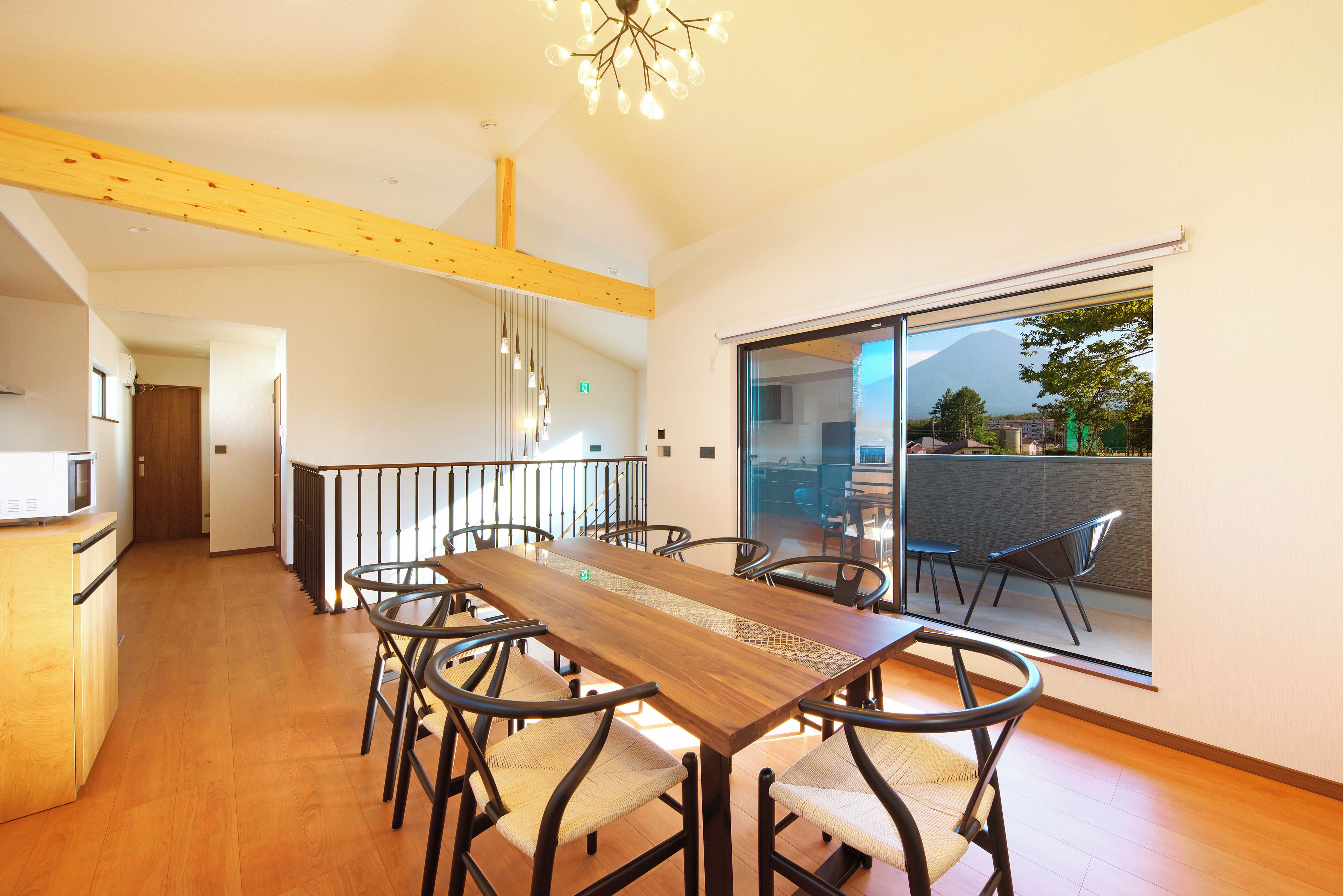 A modern dining area with a wooden table and chairs, a kitchen in the background, and a view of a mountain through a sliding glass door.
