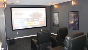 A home theater room with a large screen displaying a movie poster, black leather reclining chairs, and wall-mounted speakers.
