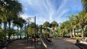 A playground with a slide and climbing structures, surrounded by palm trees and a clear sky. - 2 bed, 2 bath condo close to Disney and other theme parks Orlando (Kissimmee)