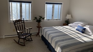 A bedroom with a bed, a rocking chair, two windows with ocean views, a ceiling fan, and a nightstand with a lamp. - Lakeside Life on Keuka's Water's Edge (Hammondsport)