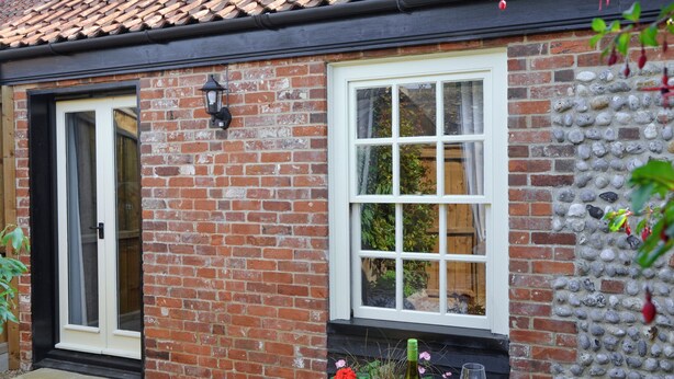 A brick house with a white window and door, a table with a bottle and glasses, and a stone wall.