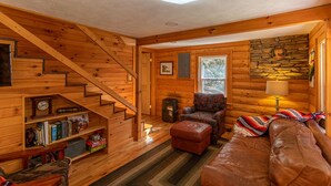 A wooden cabin interior with a staircase, a sofa, a patterned armchair, a lamp, and a stone wall. - Hidden Cabin on Old Field Creek Quaint cottage near Boone with creek frontage (Fleetwood)