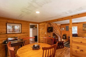 A cozy cabin interior with a wooden dining table, chairs, a kitchen area with a refrigerator, and a staircase leading to another room.