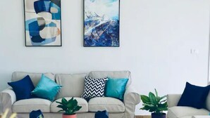 Una sala de estar con un sofá color beige, cojines azules y turquesa, una mesa de centro blanca y dos plantas en macetas.