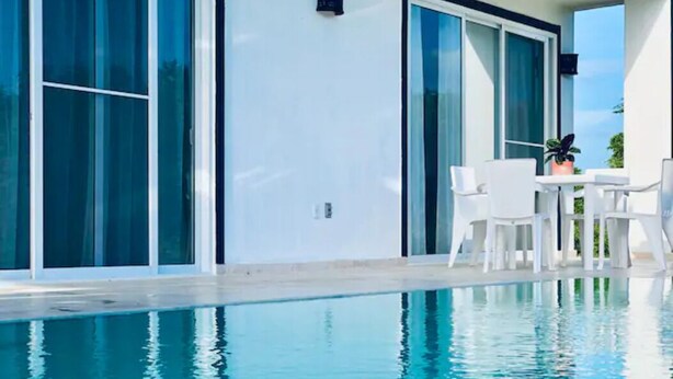 Un área de piscina moderna con agua azul cristalina, mobiliario exterior blanco y amplias puertas correderas de vidrio.