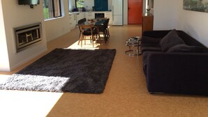 A modern living room with a dark grey sofa, a glass coffee table, and a fireplace. There is a dining area with a table and chairs, and a kitchen in the background. - House wth the Orange Door (Tasman)