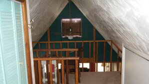 Una habitación con techo inclinado, vigas de madera, ventilador de techo y una ventana con persianas.