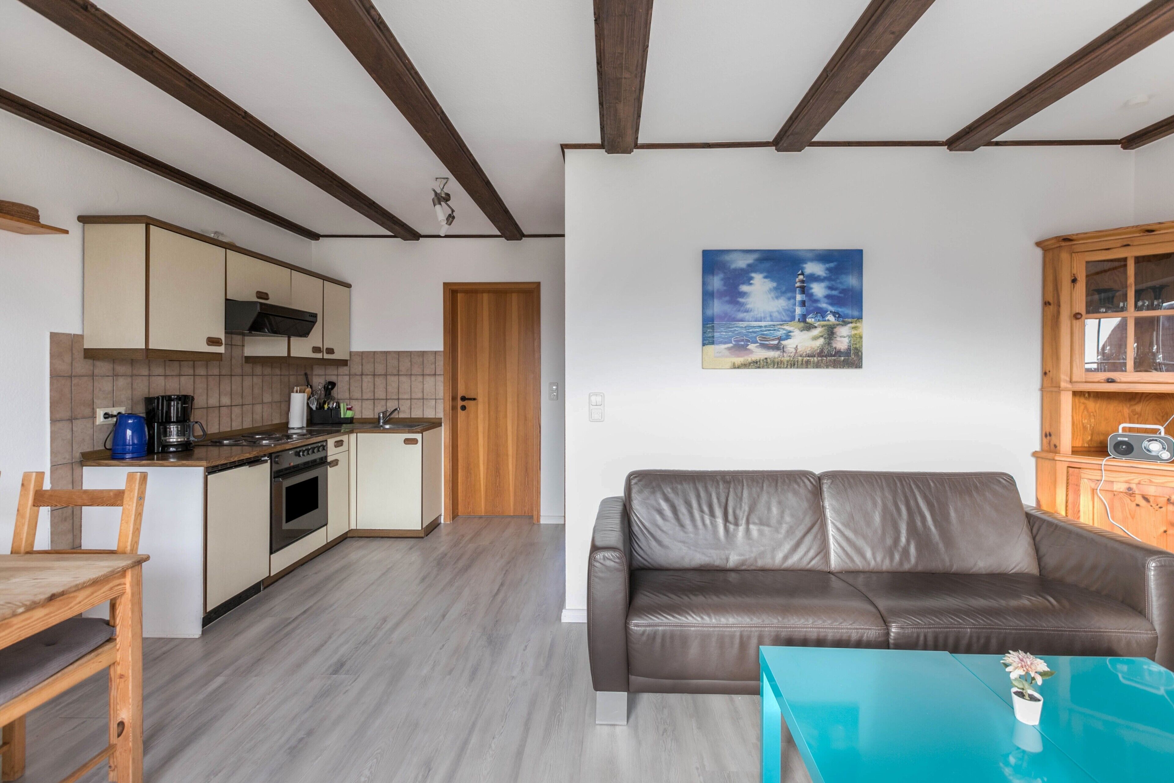 A modern kitchen with wooden beams, a dining area with a table and chairs, a living room with a sofa, and a painting of a lighthouse on the wall.