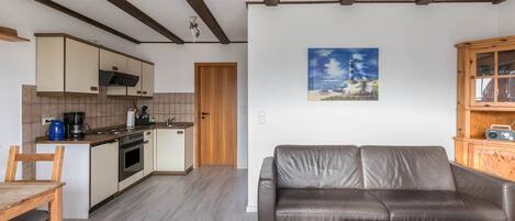 A modern kitchen with wooden beams, a dining area with a table and chairs, a living room with a sofa, and a painting of a lighthouse on the wall.