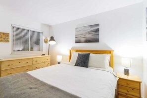 Een slaapkamer met een bed, nachtkastjes, een raam met jaloezieën, een aan de muur bevestigd schilderij en een houten dressoir.