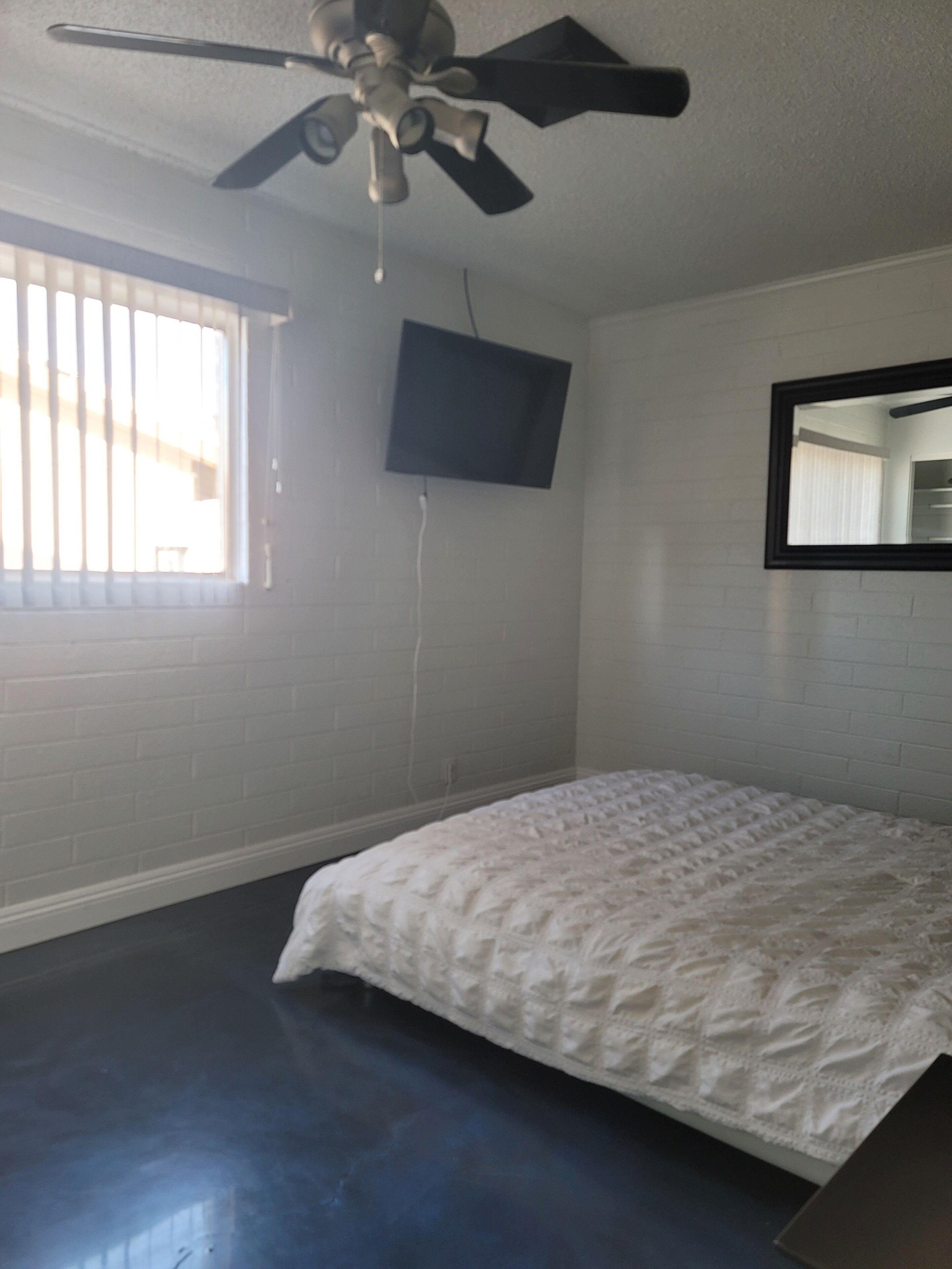 Een slaapkamer met een plafondventilator, een aan de muur bevestigde televisie, een raam met zonwering en een bed met een witte dekbedovertrek.