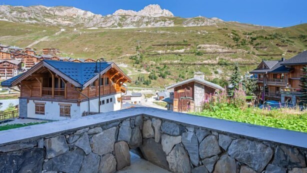 Een berghut met stenen muren en houten chalets, gelegen voor een achtergrond van rotsachtige heuvels en heldere lucht.