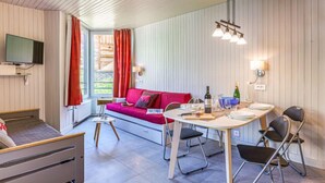 Een moderne woonkamer met een rode bank, een eettafel met stoelen, een flatscreen televisie aan de muur en een raam met gordijnen.
