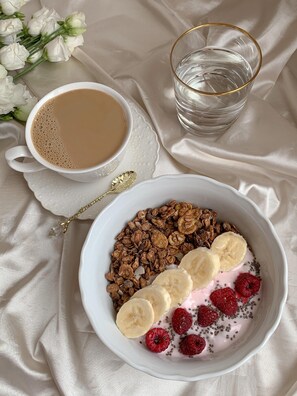 Een kom met cornflakes, bananen en frambozen, een kop koffie en een glas water op een witte tafelkleed.