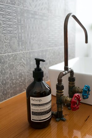 Een fles Aesop handzeepgel naast een wastafelkraan met een rode hendel.