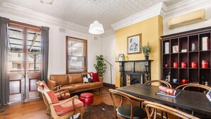 A living room with a fireplace, a dining table with chairs, a sofa, and a large window with blinds. - William Arnott Boutique Hotel (Morpeth)