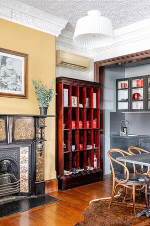 Een kamer met een open haard, een boekenkast, een eethoek met stoelen en een keuken met kasten.