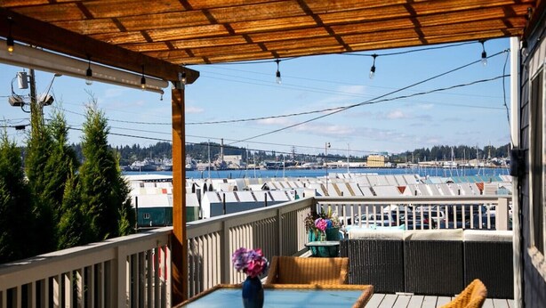 A covered outdoor area with a glass table and wicker furniture, overlooking a marina with boats and a distant cityscape. - The BayAway 4 BR Waterview Home in DT Port Orchard (Port Orchard)
