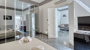 Una habitación de hotel moderna con una cama, un televisor, una zona de estar con un sofá y una silla, y un espejo grande.