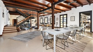 A spacious dining area with a long white table and wireframe chairs, wooden beams on the ceiling, and a staircase leading to another level. - Splendid Former Chapel With Pool and hot tub (Port Mahon)
