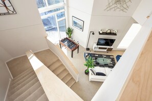 Un vaste espace de vie comprenant un canapé, une table basse et une table de baby-foot. Un escalier mène à un autre niveau.