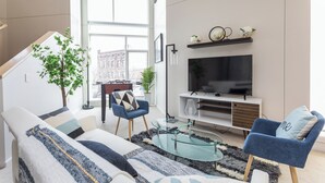 Ein modernes Wohnzimmer mit einer Couch, Sesseln, einem Glastisch, einem Fernseher und einer Treppe, die zu einer oberen Etage führt.
