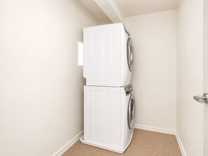 A laundry room with a stacked washer and dryer, beige walls, and a carpeted floor. - Modern Penthouse on Mass By CozySuites (Indianapolis)